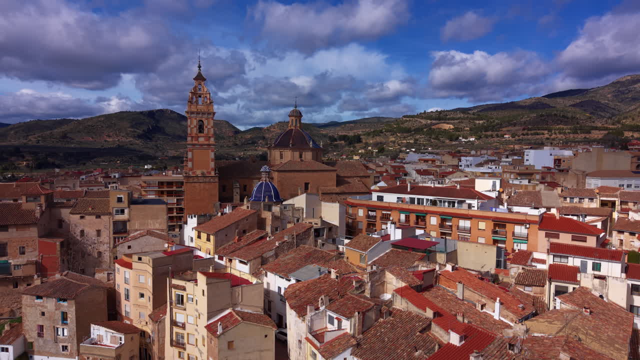 Vista elevada de Chelva, España, mostrando tejados de terracota, una torre de iglesia barroca y una cúpula de azulejos azules, rodeada de colinas verdes bajo un cielo nublado