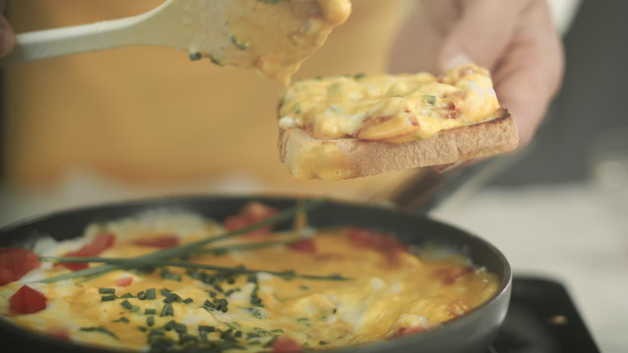 chef poniendo huevos revueltos veganos con tomates recién cocidos y rodajas de cebollino de una sartén sobre una tostada con una cuchara de madera y espolvorea cebolletas frescas sobre ella