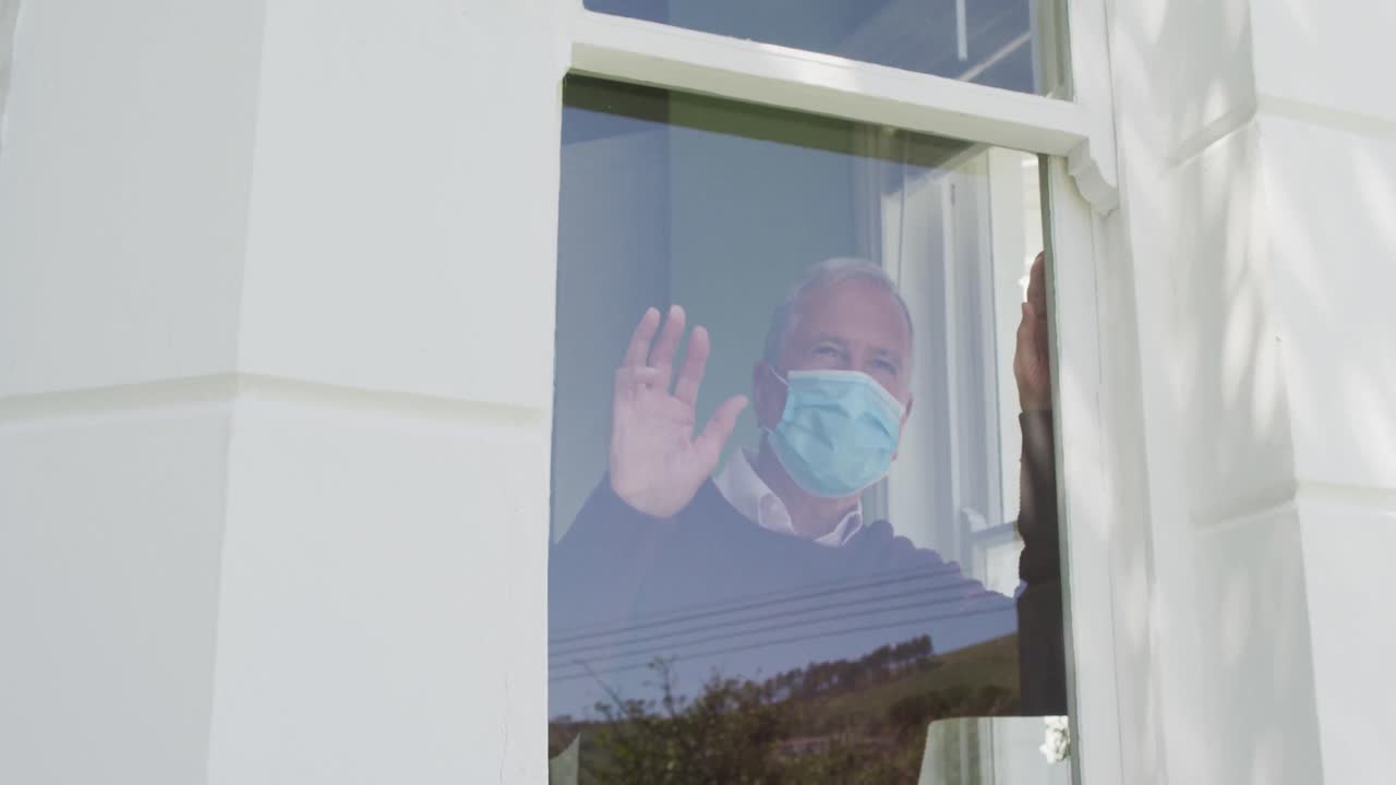 집에 있는 고위 백인 남자가 얼굴 마스크를 쓰고 창문 앞에 서서 손을 흔들고 있습니다.