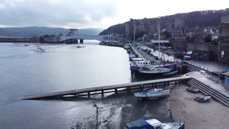 Idyllic-Conwy-castle-and-harbour-fishing-town-boats-on-coastal-waterfront-aerial-slowly-push-in-low-angle