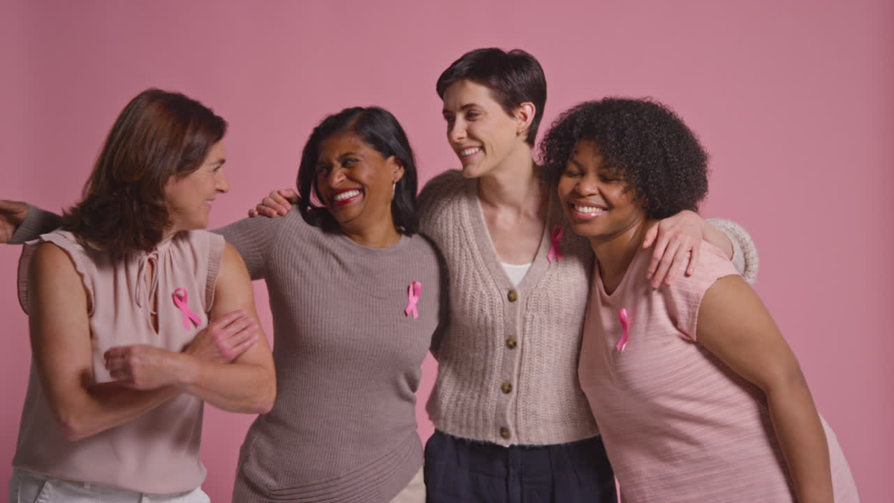 retrato de estudio de un grupo multirracial de mujeres sonrientes de diferentes edades que llevan ropa rosa y cintas de concienciación sobre el cáncer de mama abrazándose contra un fondo rosa