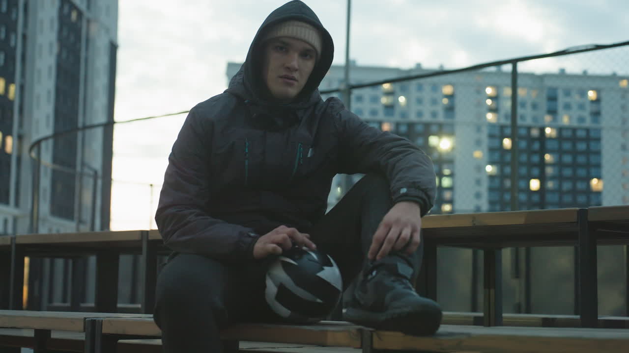 Man in hoodie sitting on bench spinning soccer ball during outdoor training session in urban sport arena, surrounded by modern residential buildings with glowing evening lights