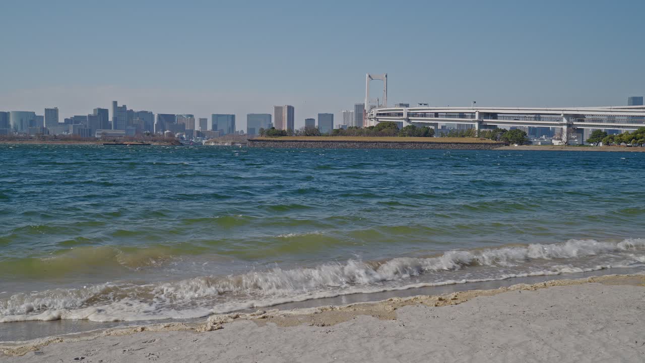 Onde turchesi si infrangono sulla spiaggia sabbiosa del Parco Marino di Odaiba, con il Rainbow Bridge e lo skyline di Tokyo che si estendono all'orizzonte, ripreso con un'inquadratura ampia e statica sotto un cielo azzurro e limpido.