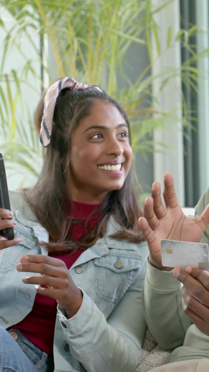 Video verticale: Donna sorridente in jeans che mostra il telefono sul divano, uomo con maglione color salvia che festeggia una carta