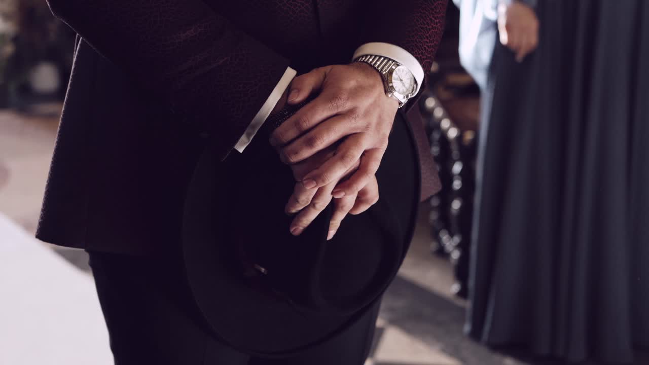 Close up of a man's hands holding a black hat, wearing a watch and formal suit