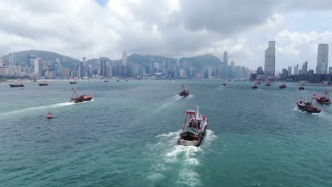 Convoy-of-local-Fishing-boats-causing-in-Hong-Kong-Victoria-bay,-with-city-skyline-in-the-horizon,-Aerial-view