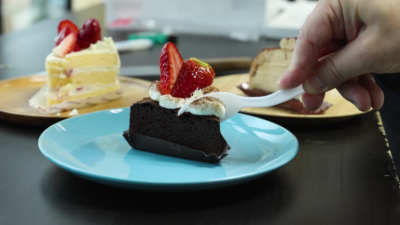 Hand uses spoon to scoop chocolate cake topped with cream and strawberries on blue plate