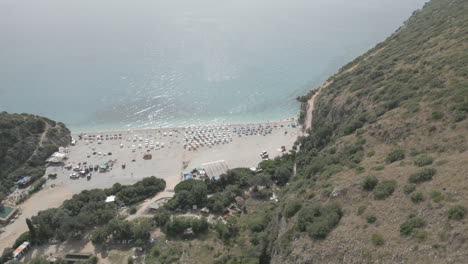 Drone-Disparó-Sobre-La-Playa-De-Gjipe-Albania-Con-Turistas-En-La-Playa-Y-Agua-Azul-Clara-Cerca-De-Las-Montañas-Y-El-Mar-En-Un-Día-Soleado-Y-Brillante-Y-Naturaleza-Verde-Alrededor-Con-Tumbonas-Y-Toallas