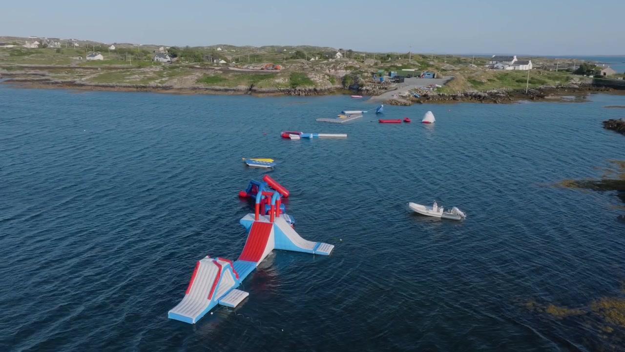 Colorful inflatable water park modules float offshore near a small boat and rocky Lettermullan coastline under clear summer skies.