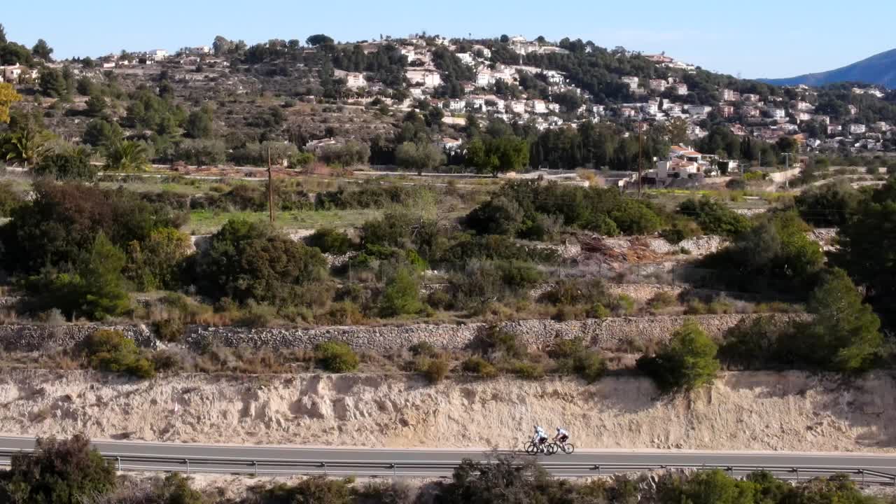 Panning drone footage of three team cyclists riding along a rural tarmac road in a valley, on a hot day in Calpe, Spain with trees and traditional whitewashed buildings in the background