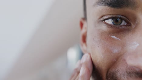 primer plano de un hombre biracial mirándose en el espejo aplicando crema facial debajo de los ojos en el baño, cámara lenta