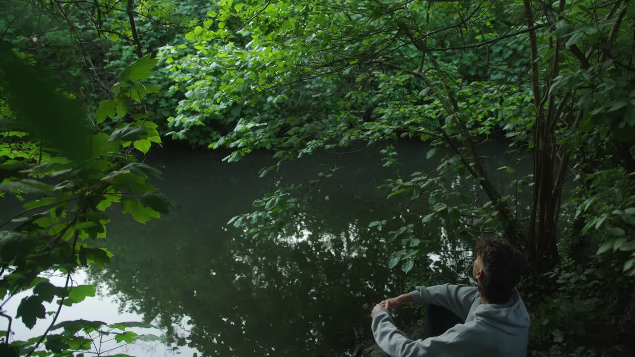 Man rests by a calm forest river surrounded by thick green foliage