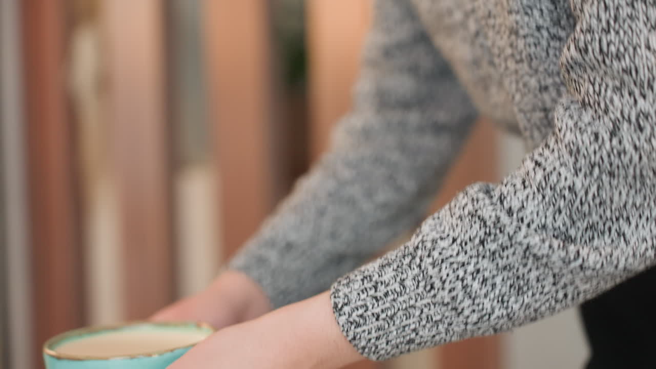 Young woman gently walking toward table holding hot coffee in hands carefully preparing to place cup down and sit relaxed moment cozy indoor ambiance textured sweater cup close view