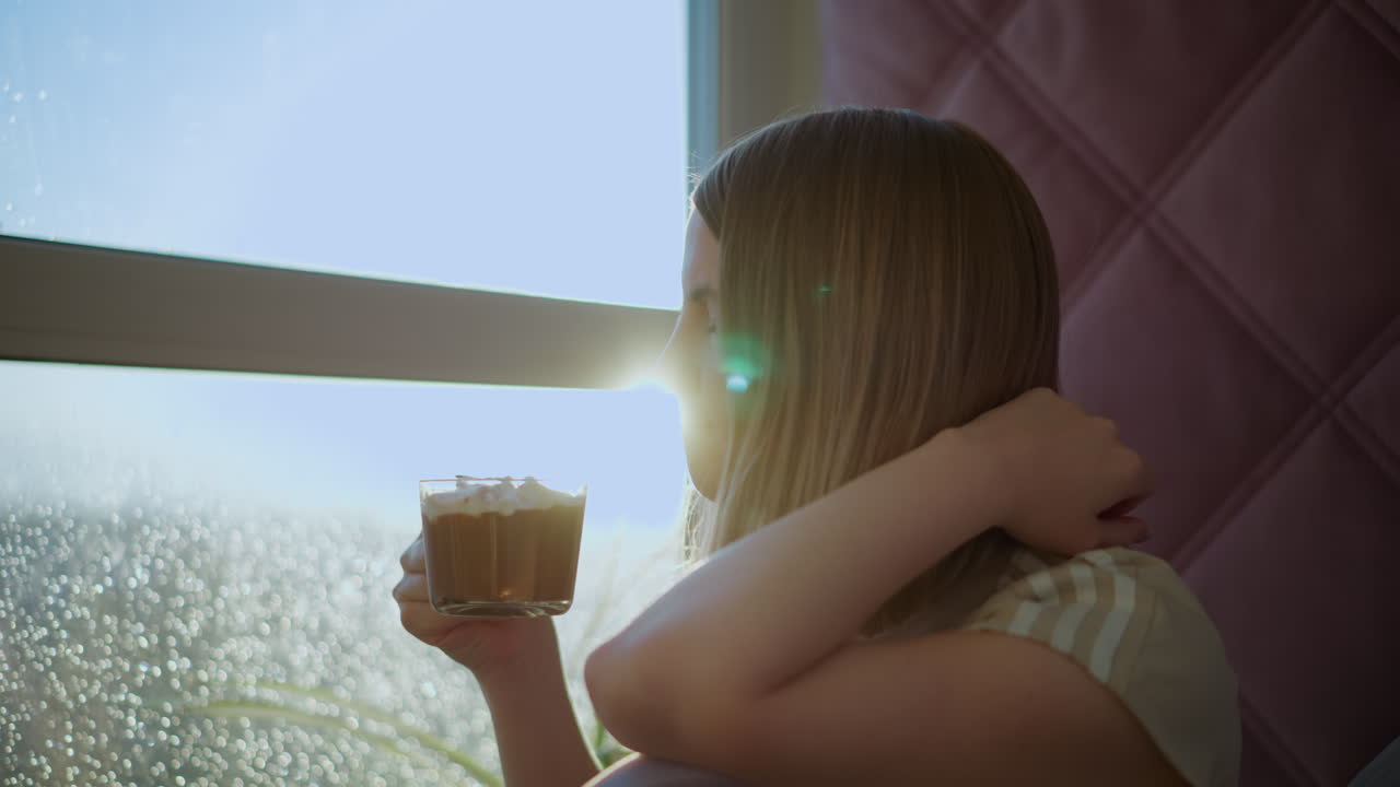 Young woman sitting near window with bright sunlight shining through glass, holding cup with whipped cream topping, touching hair, enjoying warm peaceful moment with soft bokeh background and cozy atmosphere