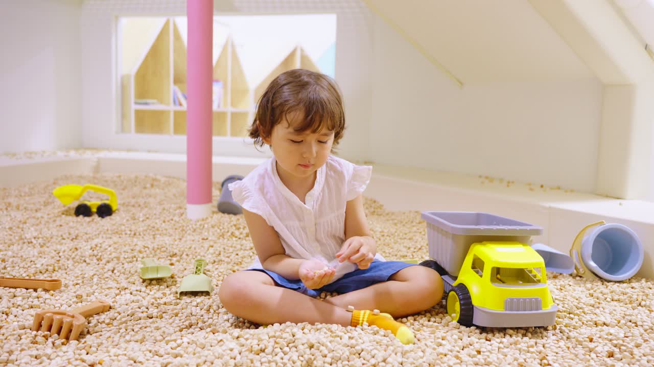 Cute 5 year old girl in high heels sitting indoors in sandbox filled with beige granules, stacking them in her hand