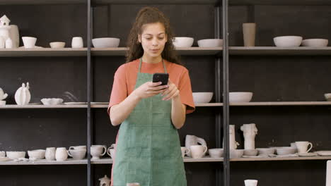 woman in apron using phone and taking photos of handmade ceramic pieces on table in the pottery shop