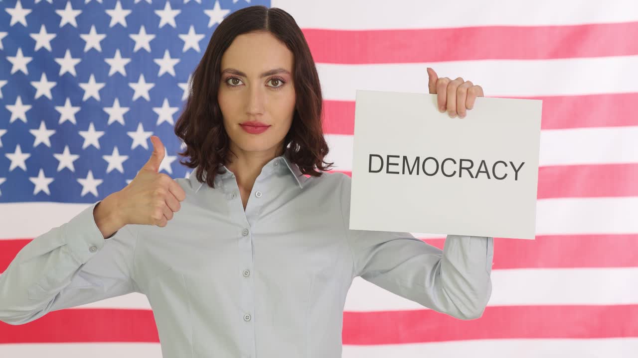 Woman holds 'DEMOCRACY' sign in front of an American flag and gives a thumbs-up