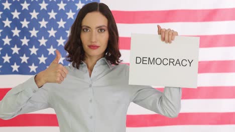 woman holds 'democracy' sign in front of an american flag and gives a thumbs-up