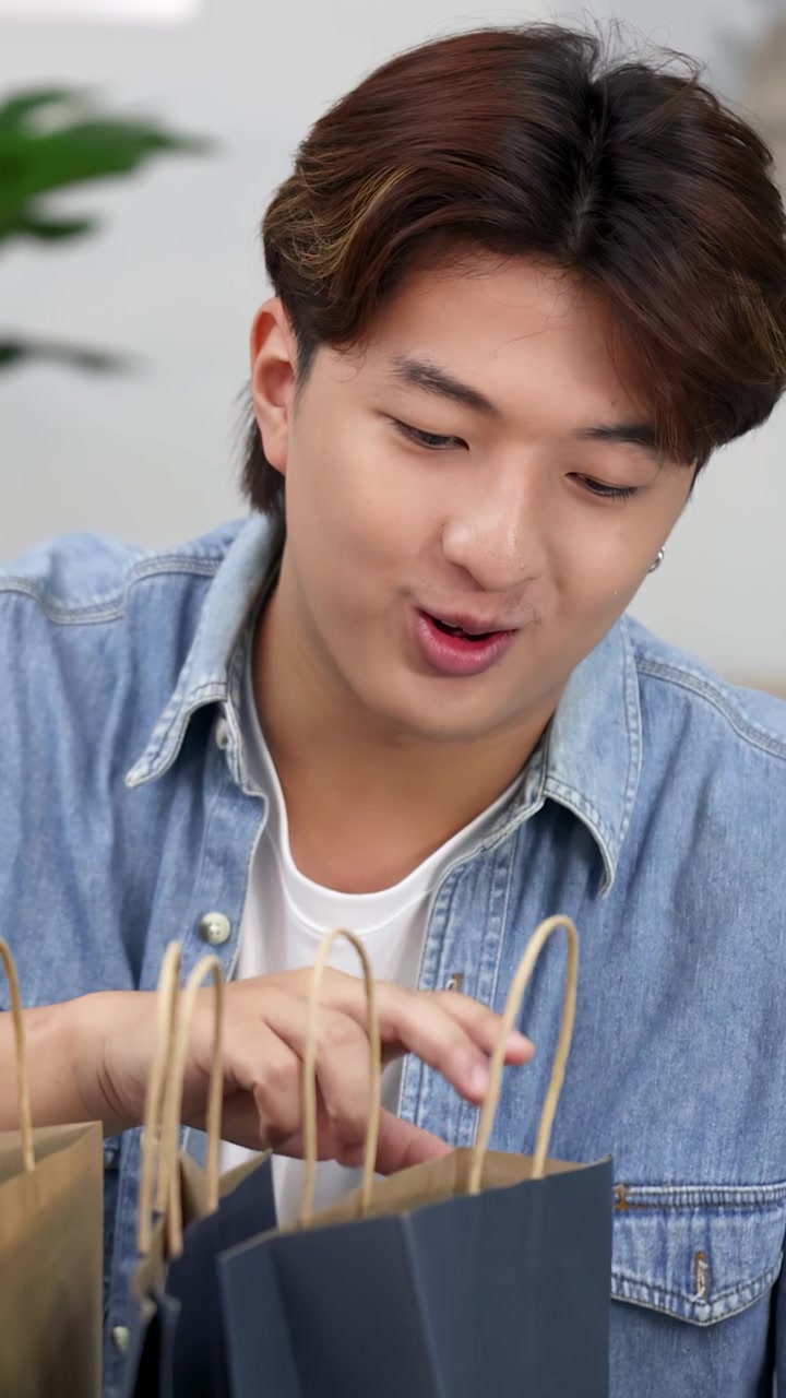 明るいリビングルームで、陽気なアジア人男性がいくつかの紙製ショッピングバッグを調べている。