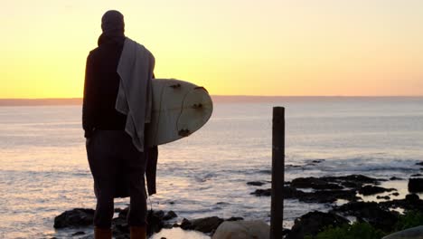 Vista-Trasera-De-Un-Hombre-Adulto-De-Mediana-Edad-Con-Una-Tabla-De-Surf-Parada-Cerca-De-La-Playa-4k