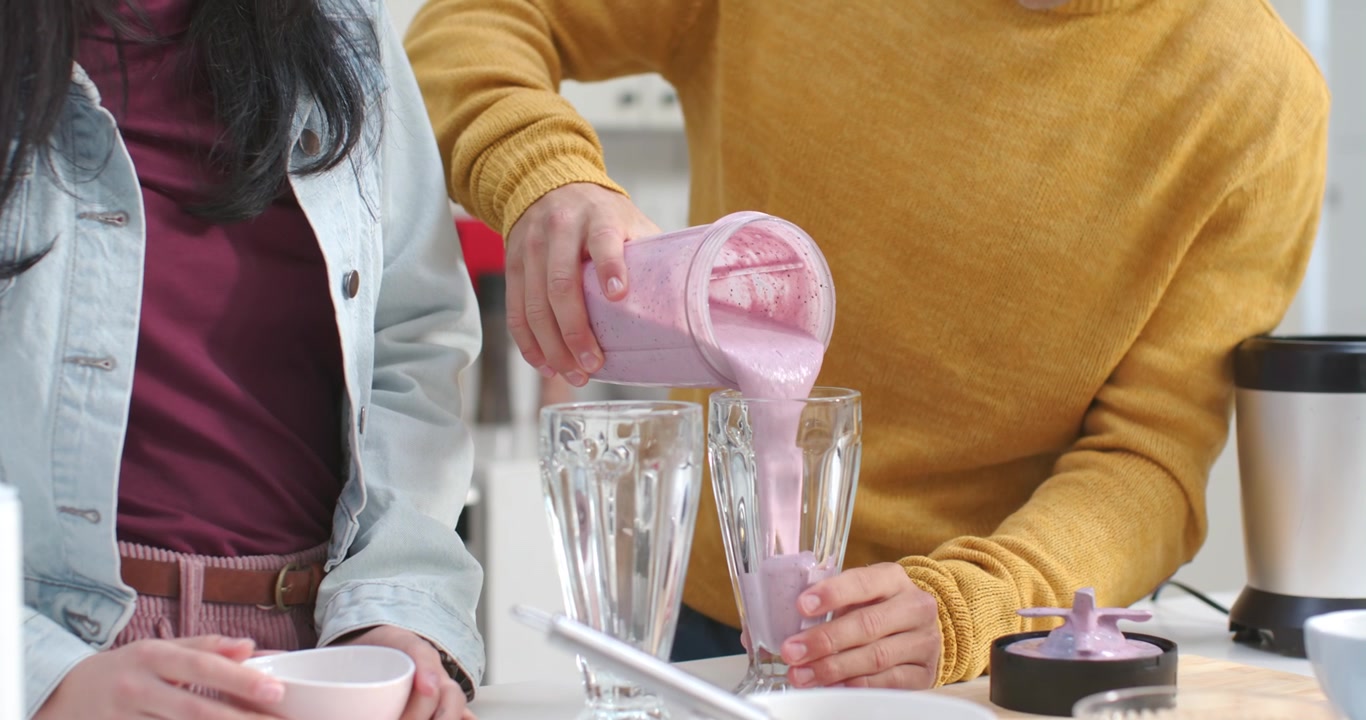 Una persona con un maglione color senape sta versando un frullato rosa in due bicchieri sul bancone per servirli.