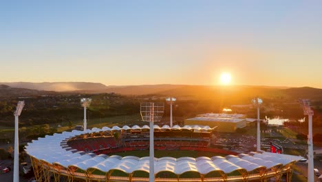 drone footage circles a large illuminated stadium at sunset, revealing surrounding landscape, river, and event lighting in a warm, golden hour ambiance