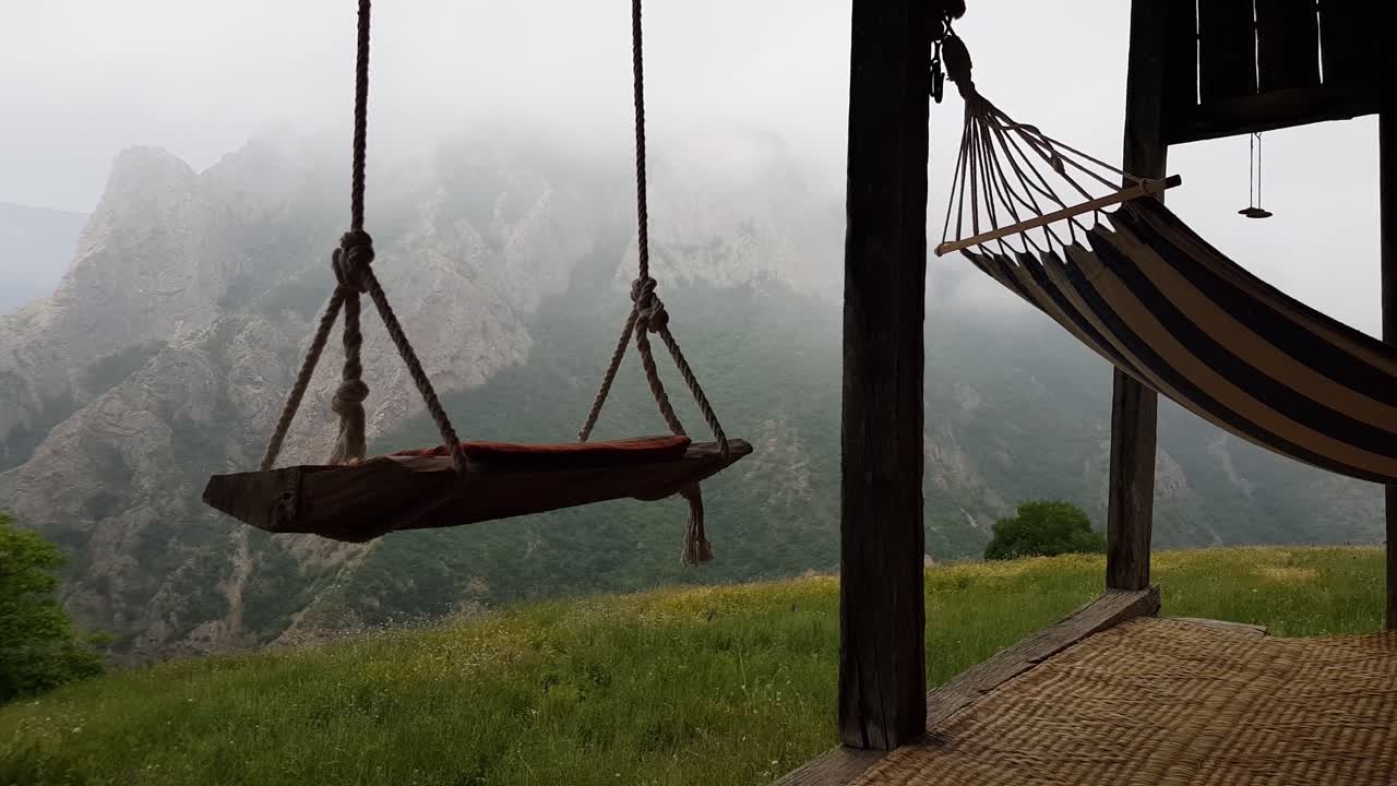 columpio de cuerda de madera y hamaca de algodón relajante en una hermosa terraza de cabaña con alfombra tradicional y prado verde y montaña rocosa en el paisaje de niebla