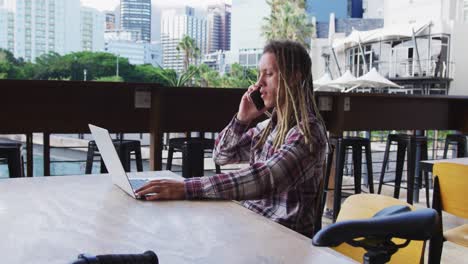 Mixed-race-man-with-dreadlocks-sitting-at-table-outside-cafe-using-laptop-and-smartphone