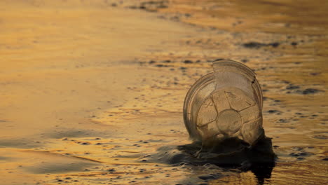 Nahaufnahme-Eines-Leeren-Plastikbechers,-Der-Am-Strand-Liegt,-Während-Das-Wasser-Am-Späten-Nachmittag-Gegen-Ihn-Spritzt