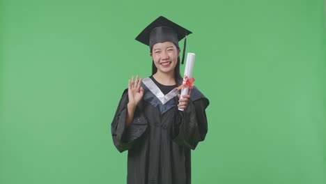 asian woman student graduates in cap and gown holding diploma, smiling, and waving her hand on the green screen background in the studio