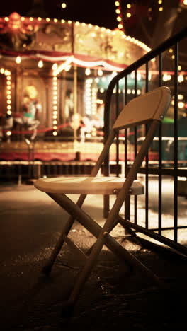 a white metal folding chair sits in front of a blurred carousel at night