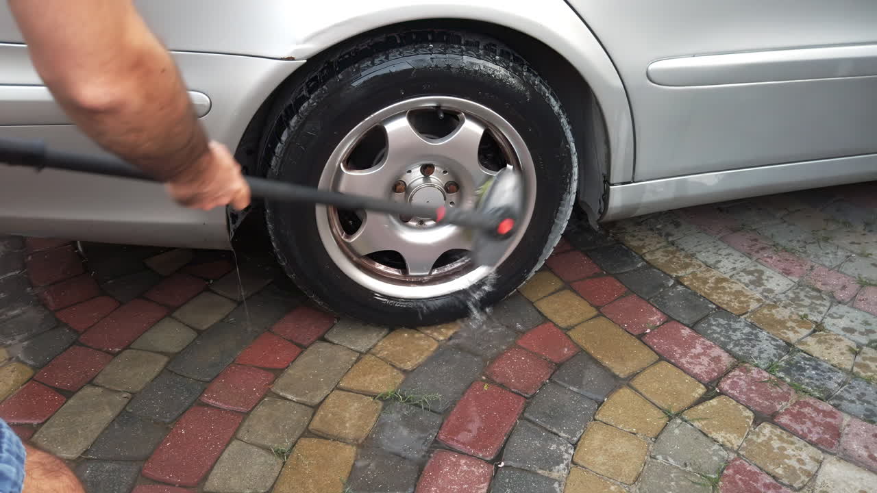 A person is using a pressure washer to clean a car tire while standing on a colorful stone pavement. The sunlight brightens the scene, showcasing the tire and pavement details