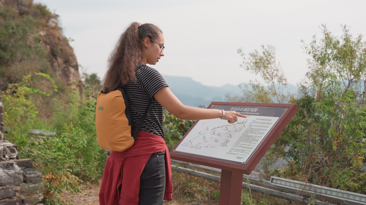 Young woman hiker reads trail map at overlook, curly hair, yellow backpack and red jacket tied around waist, pointing finger at map panel, scenic mountain vista in background, overcast sky, thoughtful