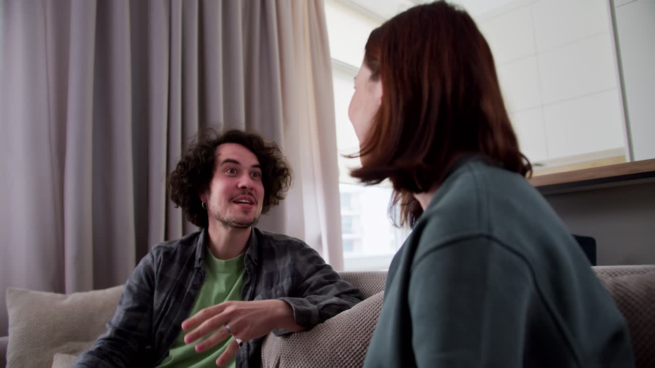 A happy brunette guy with curly hair in a checkered shirt is actively communicating with his brunette girlfriend while sitting on the couch at home