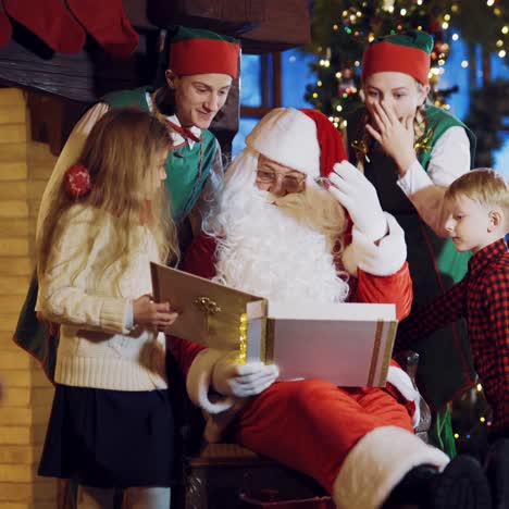 santa claus is sitting on a chair surrounded by two elves and a little boy with a girl and considering a holiday album with photos on the background of new year's decorations and the christmas tree in the room at home