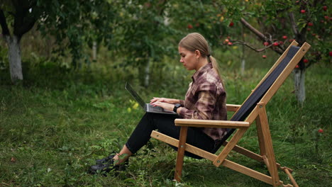 blonde young lady typing on her laptop sitting in the apple garden. woman working freelance in the nature surrounding.