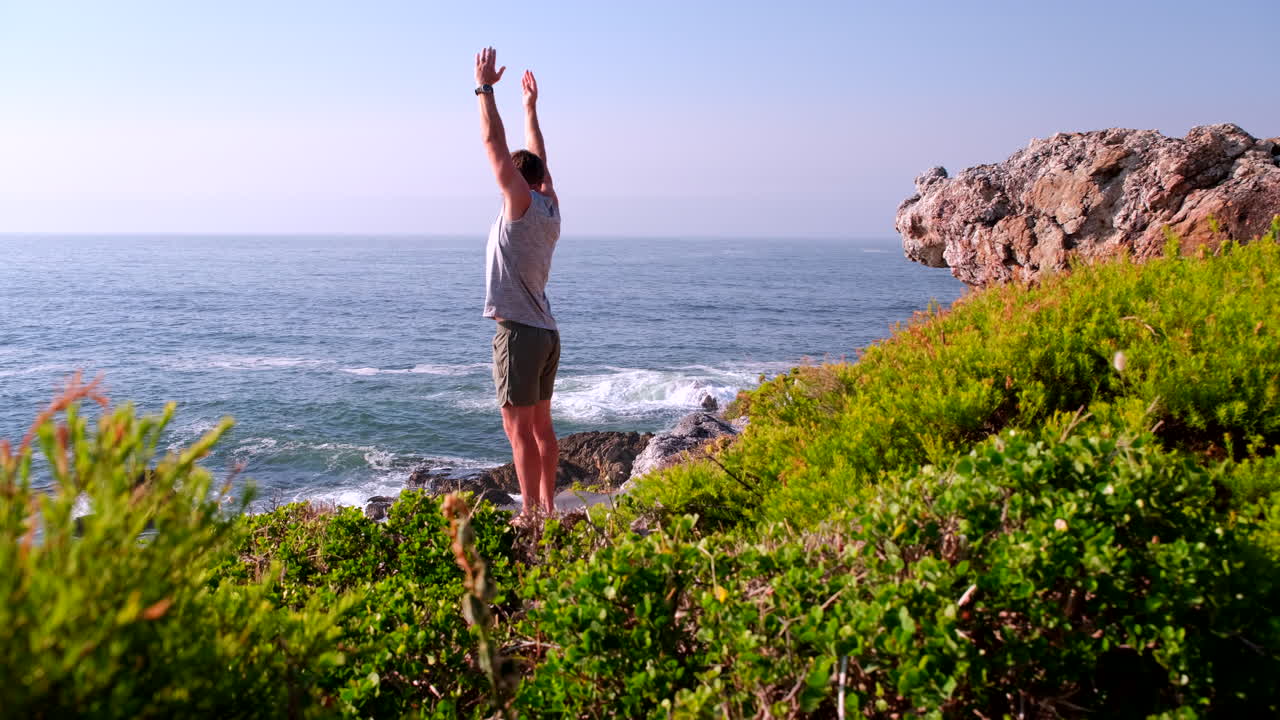 Fit male does standing side stretches with raised arms at sunrise on sea cliff