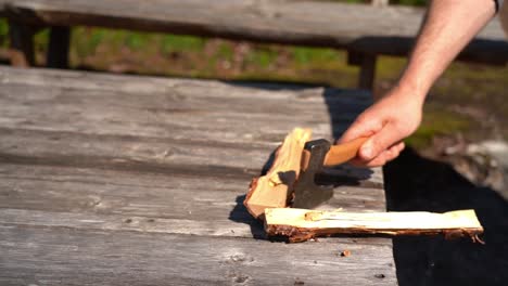 Male hand splitting wood with small hiking axe slow motion - Sunny day outdoor clip with firewood standing on wooden table before beeing split into two pieces