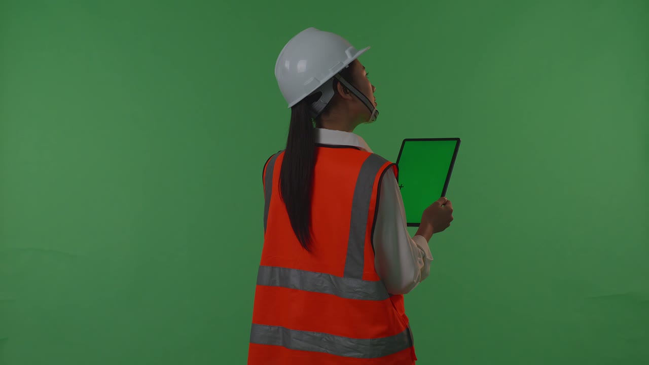 안전 헬을 입은 아시아 여성 엔지니어가 녹색 스크린 태블릿에서 작업하고, 일몰이나 해가 뜨는 동안 녹색 화면 배경 스튜디오에 서있는 동안 주변을 둘러보고 있습니다.