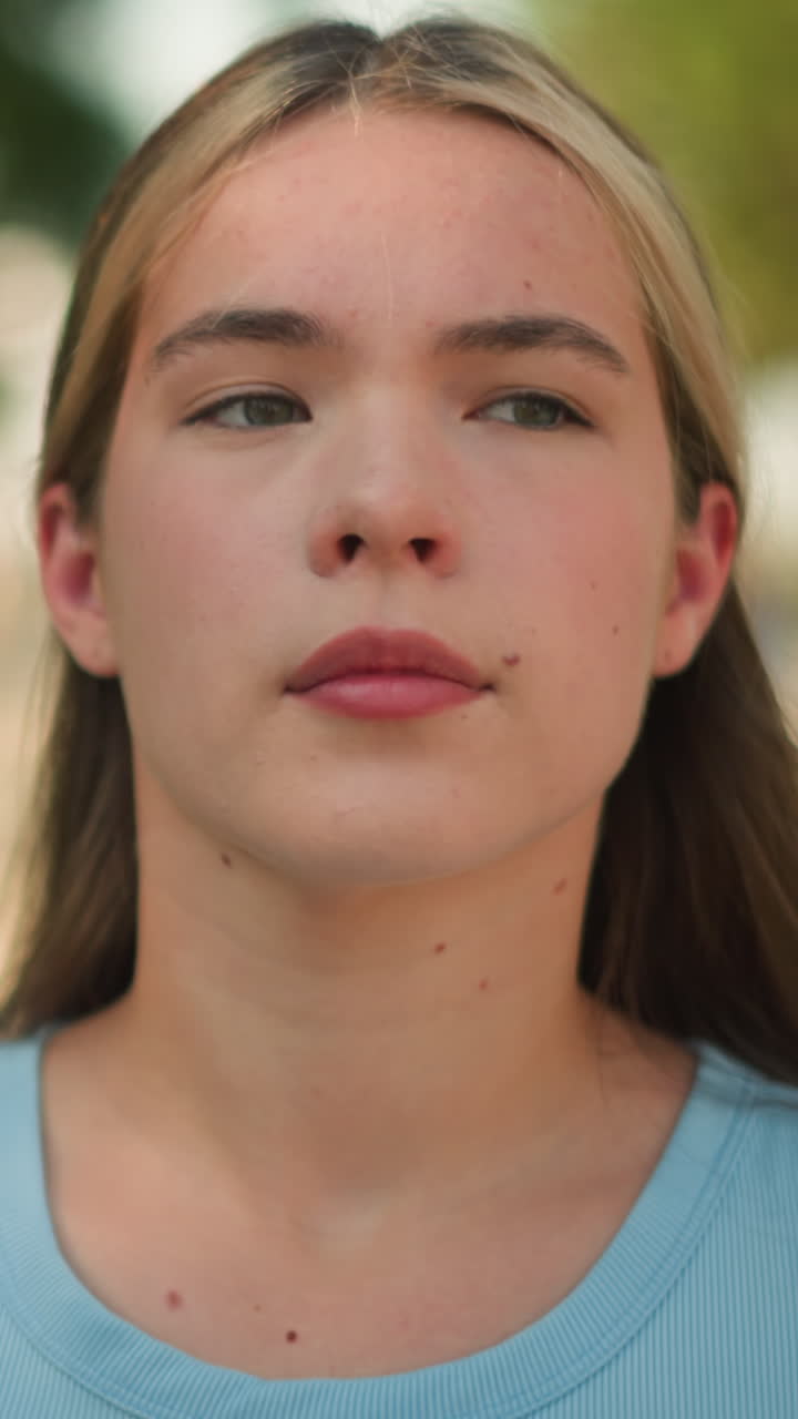 primer plano de una mujer joven con camiseta cian caminando al aire libre bajo la luz del sol, con expresión enfocada, fondo borroso con vegetación, árboles, postes de lámparas y personas
