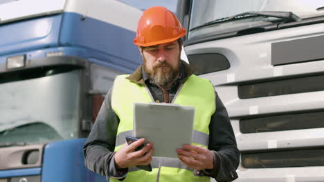 worker wearing vest and safety helmet organizing a truck fleet in a logistics park while consulting a document and holding a smartphone