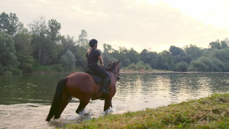 Female-horseback-rider-riding-a-horse-along-the-river-at-sunset,-side-view
