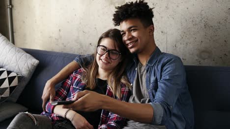 Cute-happy-couple-of-young-hipsters-smiling-and-cuddling-on-the-couch-while-trying-to-find-a-perfect-tv-channel.-Leisure-time,-enjoying-youth,-living-together.-Relationship-goals.