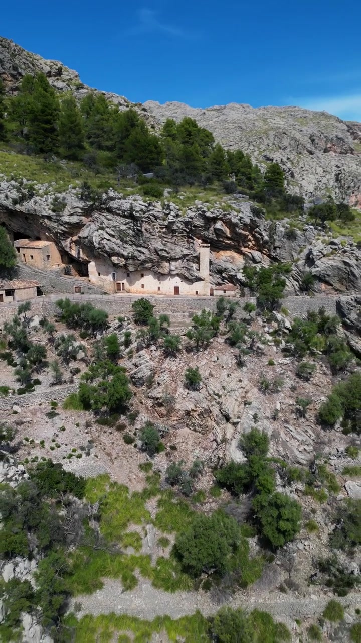 Edifici tradizionali in pietra incastonati in una scogliera sull'isola di Maiorca, Spagna, che mostrano un'architettura unica contro una vegetazione lussureggiante e un cielo azzurro limpido. Ripresa verticale ad alta velocità con allontanamento.