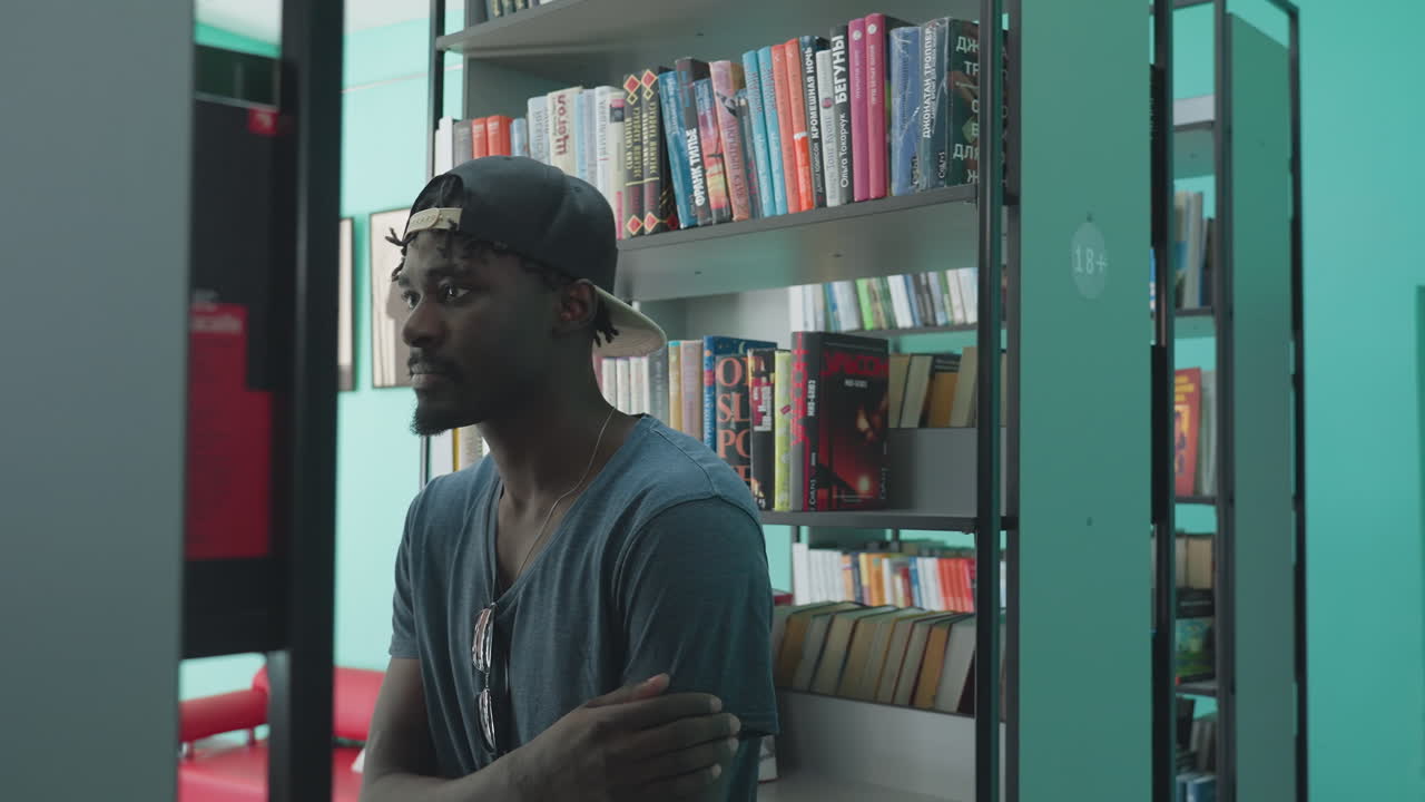 Student wearing cap and casual shirt walks into library, passing shelves filled with books and decorative items, looking thoughtful against turquoise wall in brightly lit indoor academic environment