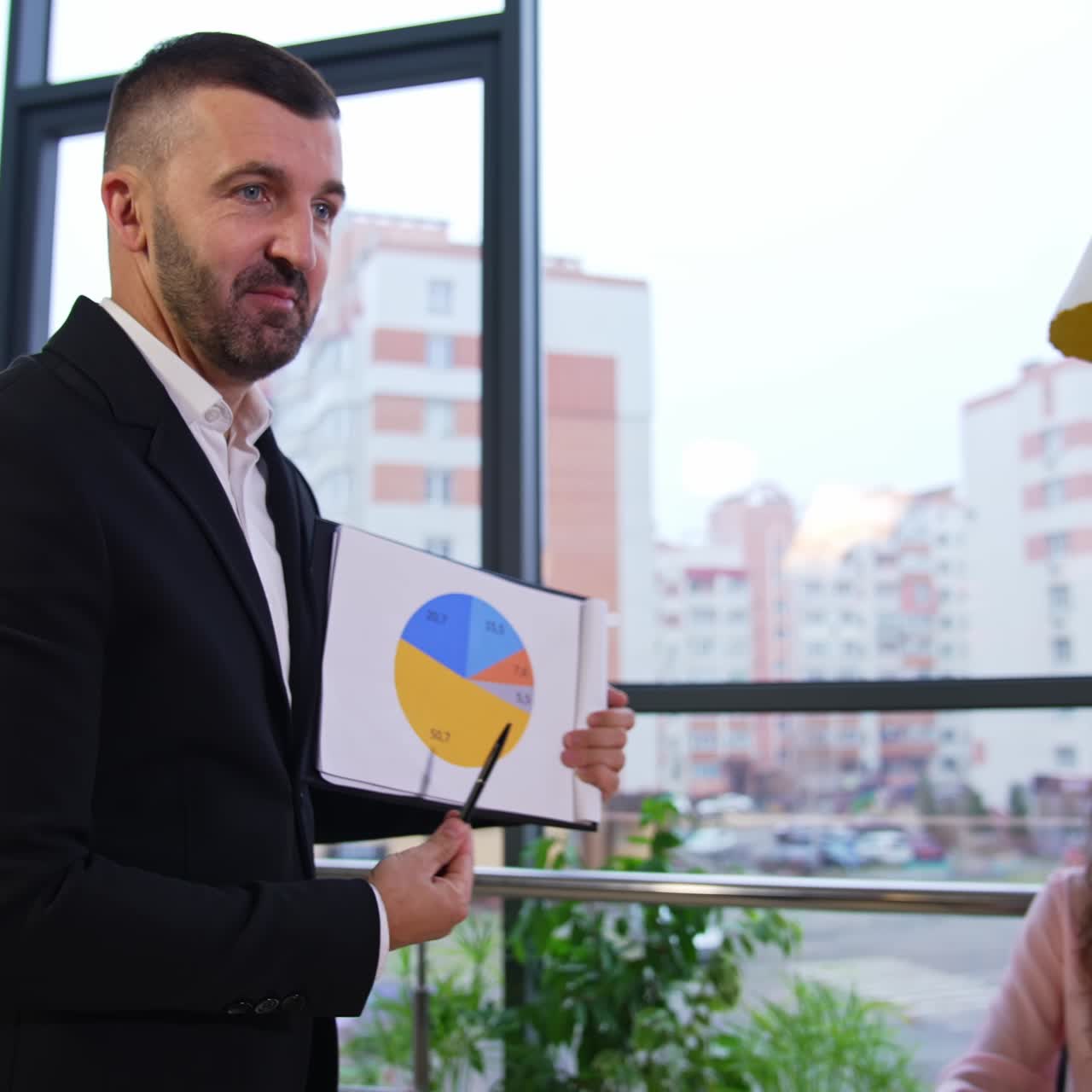 Business meeting with coffee drinking in a spacious office. Male boss holding a circle diagram and explaining it to his staff. Panoramic window and cityscape at backdrop