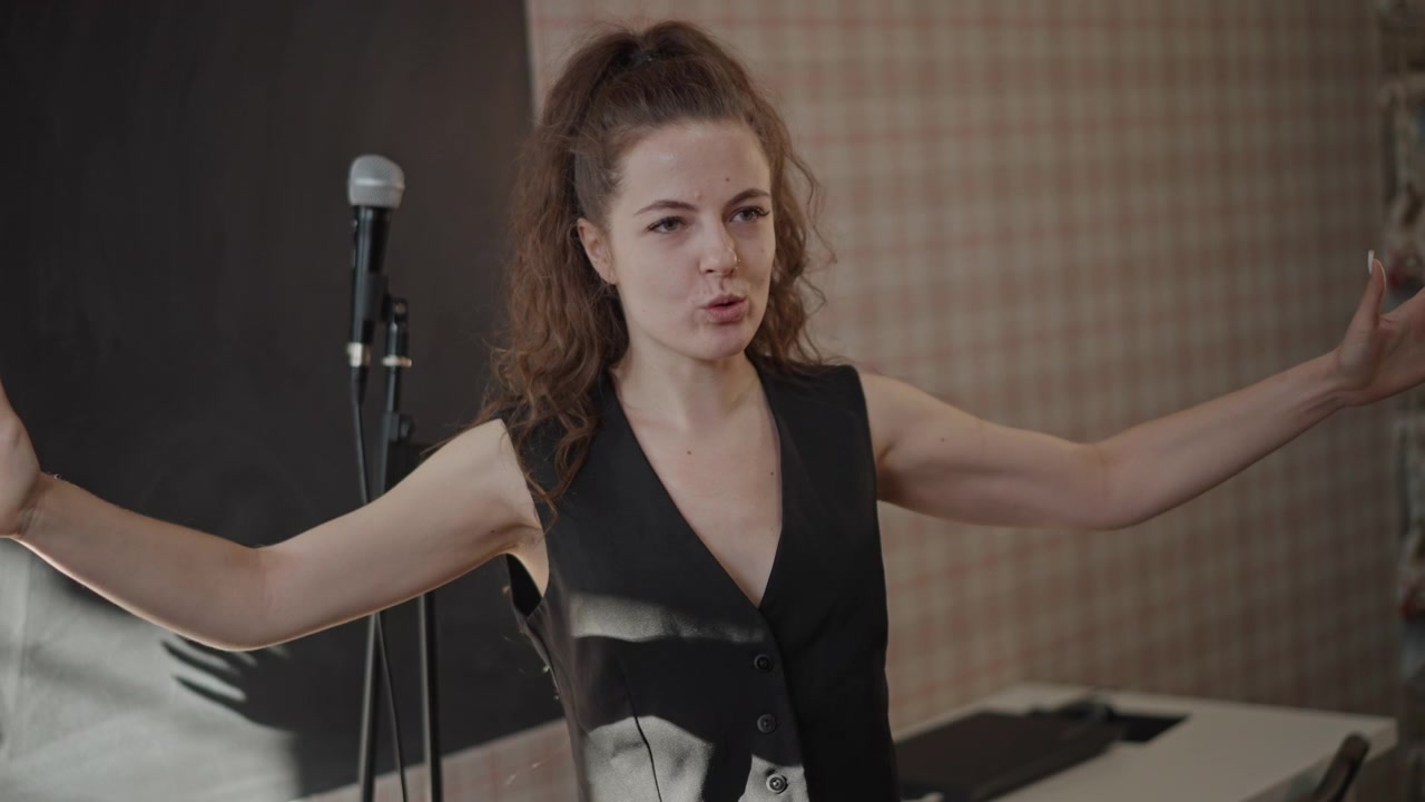 Vocal coach donna bianca che dirige una prova in uno studio accogliente, guidando gli studenti con gesti espressivi delle mani, luce solare calda, asta del microfono, attenzione al controllo del respiro e all'intonazione, lezione di musica in classe.