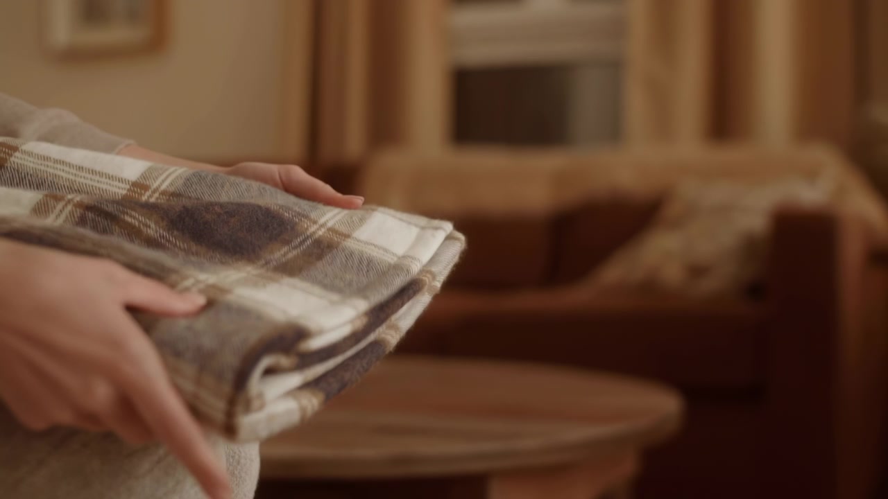 Mani che lisciando e riposizionano una coperta a quadri piegata sul bracciolo di un divano imbottito, rivelando una texture intrecciata, pieghe a strati, un arredamento neutro e morbido, luce interna calda, per un comfort domestico accogliente e rilassato.