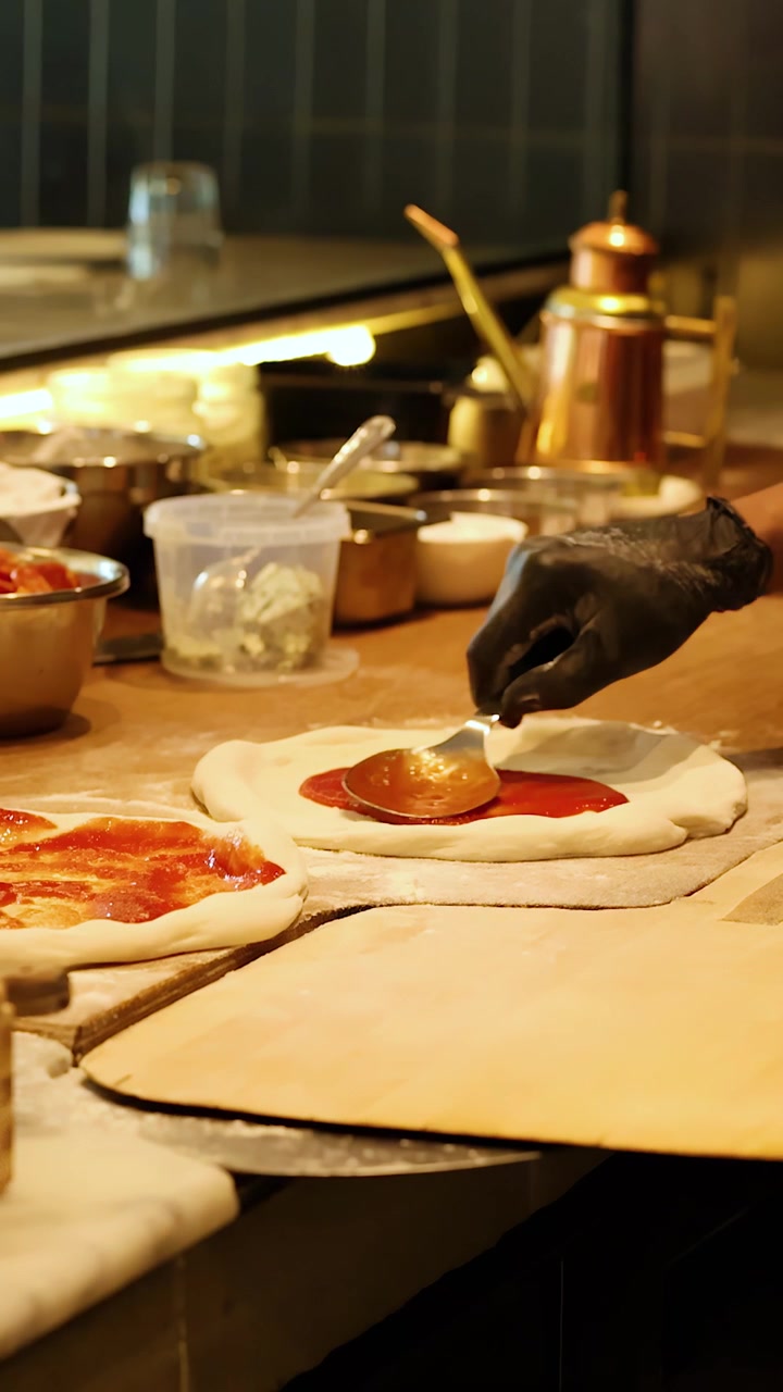 Primo piano di uno chef con guanti neri che spalma salsa di pomodoro sull'impasto usando un cucchiaio