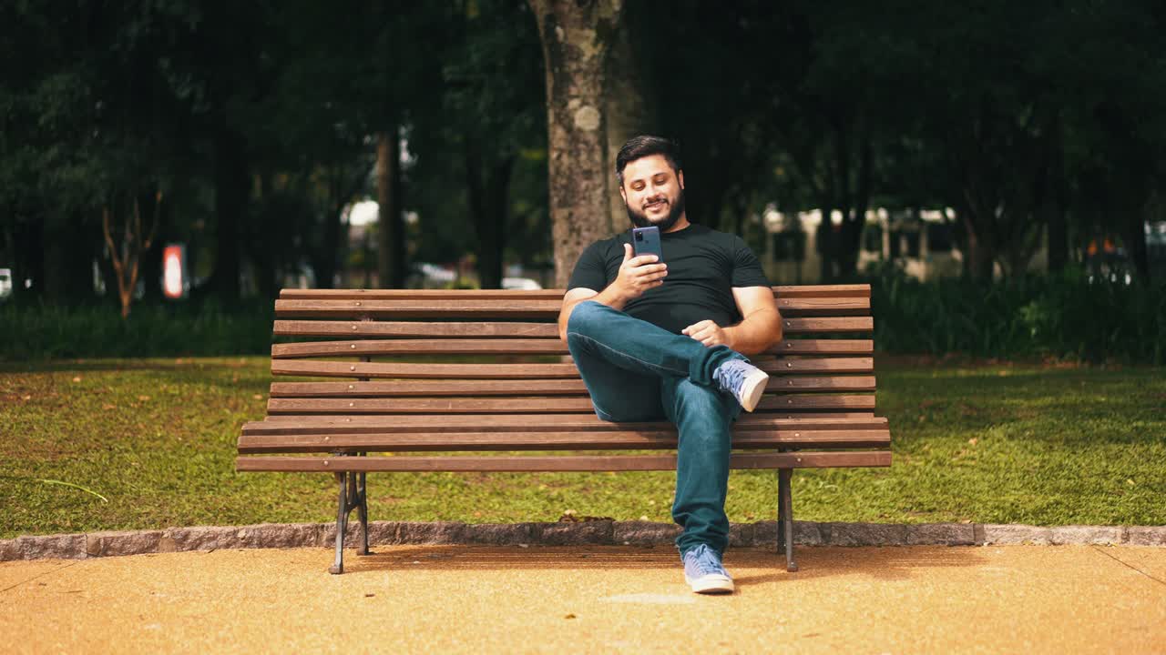 자신의 휴대 전화를 사용하여 나무 공원 벤치에 혼자 앉아 웃는 남자